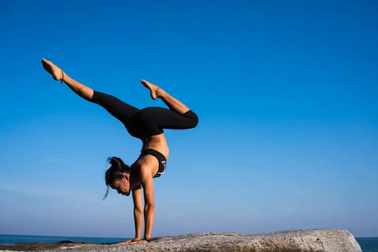 A woman wearing black athletic wear performs a handstand on a rocky surface against a clear blue sky.