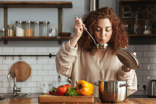 A woman with curly red hair wearing a beige sweater is blowing on a ladle filled with soup over a pot in a kitchen. These soups