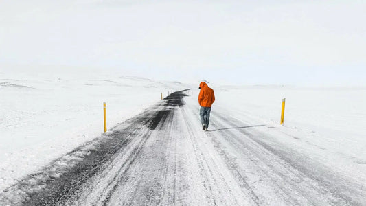 A person in an orange jacket and jeans walks down a snowy, deserted road with yellow markers on either side.