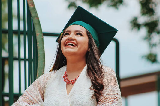 A woman in a green graduation cap and white lace dress smiles brightly while looking up, celebrating the added benefits series of her achievements.