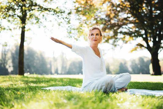 A woman sits cross-legged on a blanket in a grassy park, performing a yoga pose with one arm extended to the side.