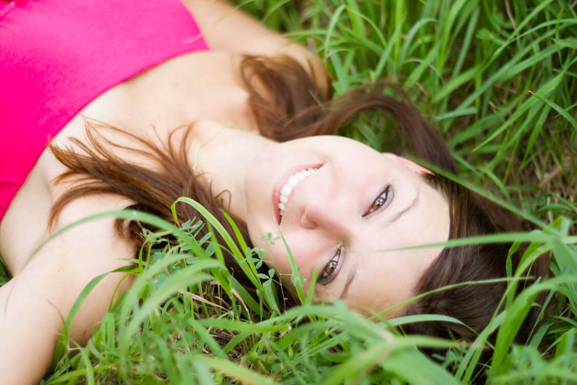 A woman is lying in a grassy field, looking up at the camera with a smile. She has long brown hair and is wearing a pink top.