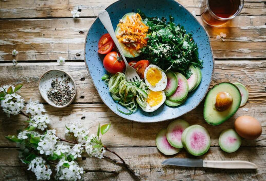A colorful, healthy meal is presented on a rustic wooden table. The blue plate contains halved boiled eggs, sliced cucumbers, avocado, tomatoes, spiralized zucchini, grated carrots, and mixed greens.