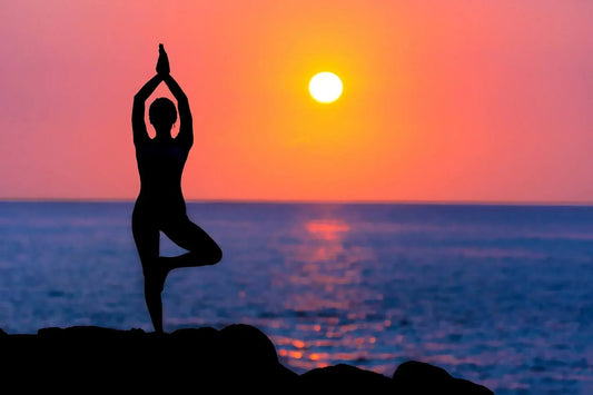 Silhouette of a person performing a yoga tree pose with arms raised above the head at sunrise or sunset.