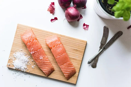 Two raw salmon fillets, rich in omega-3, rest on a wooden cutting board with a small pile of coarse sea salt beside them. In the background
