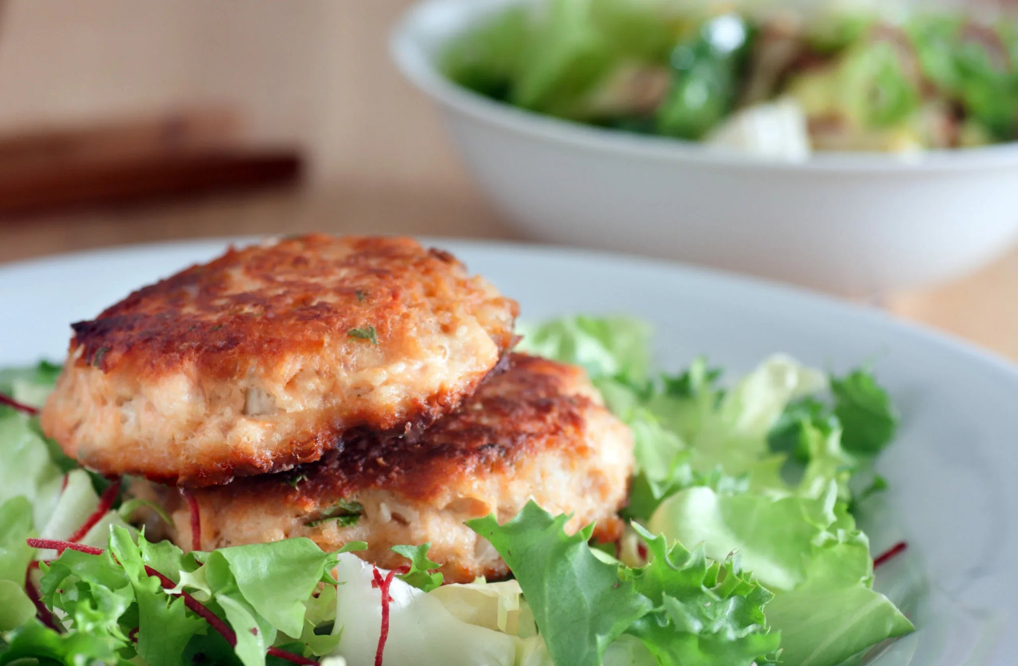 Two golden-brown salmon patties rest on a bed of fresh mixed greens, featuring lettuce, beet strips, and herbs.