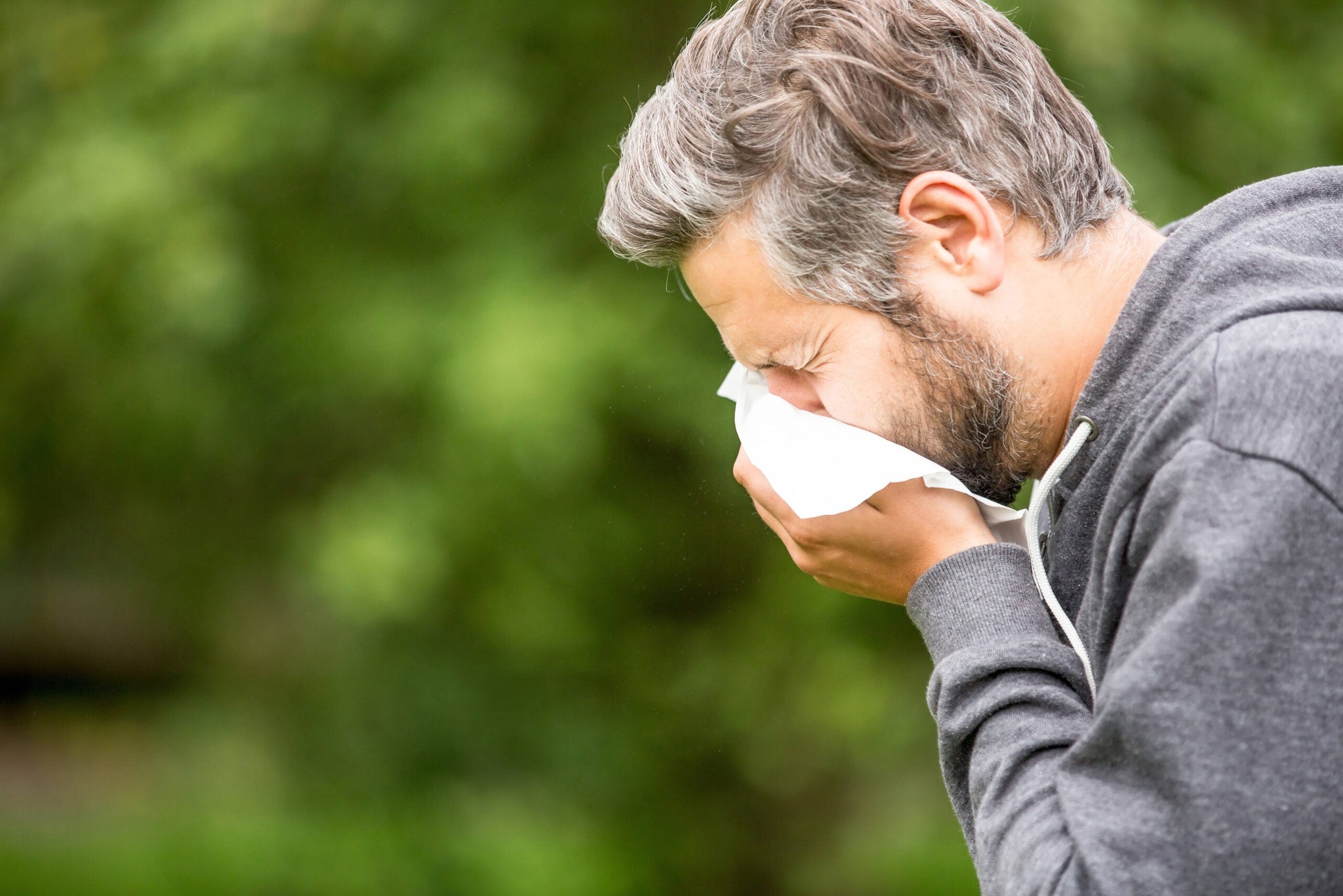 A man with gray hair and a beard is sneezing into a tissue outdoors.
