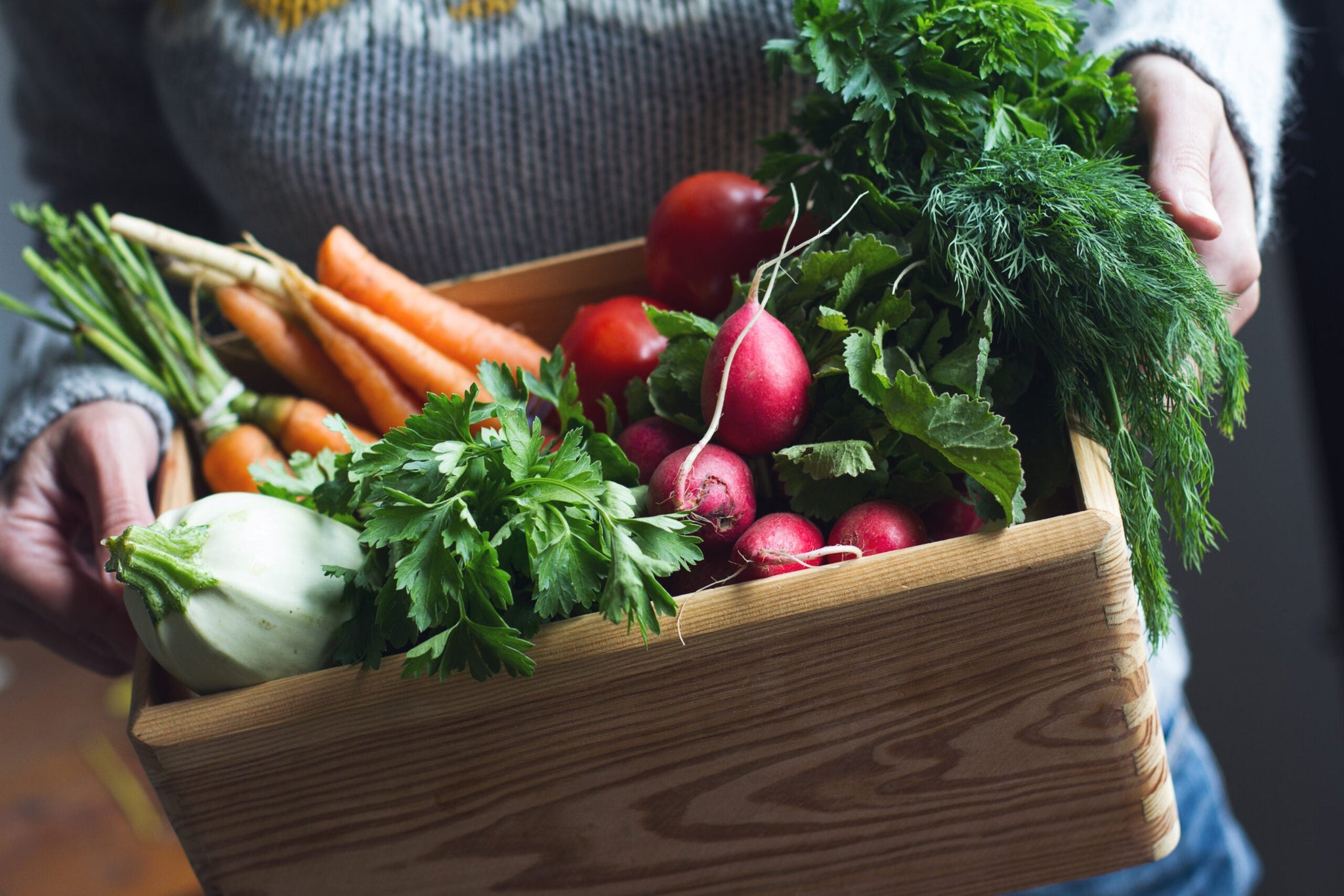 Person holding a wooden crate filled with fresh vegetables, including carrots, radishes, fennel, and leafy greens.
