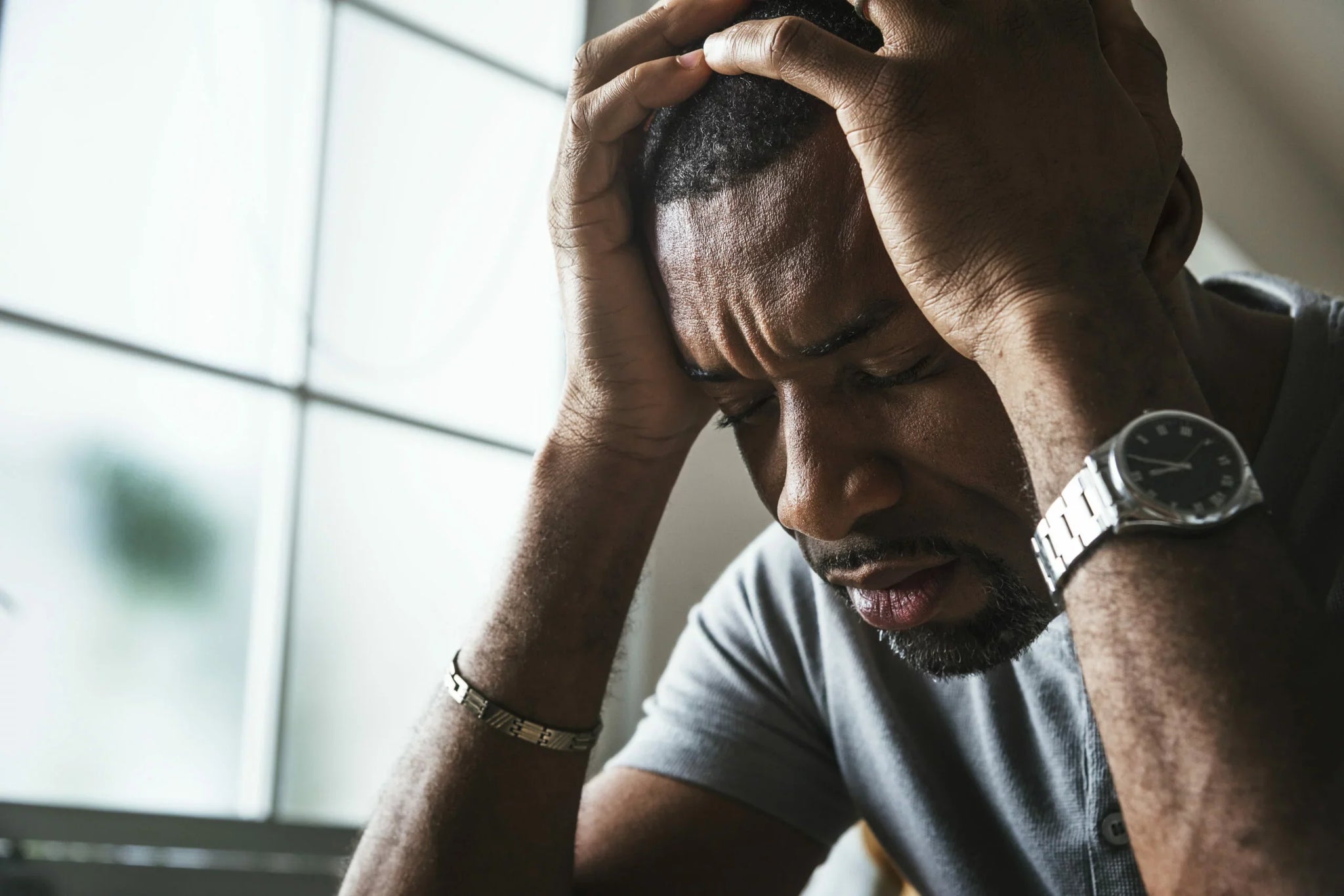 A man sits indoors, leaning his elbows on a surface with his hands clasped over his head.
