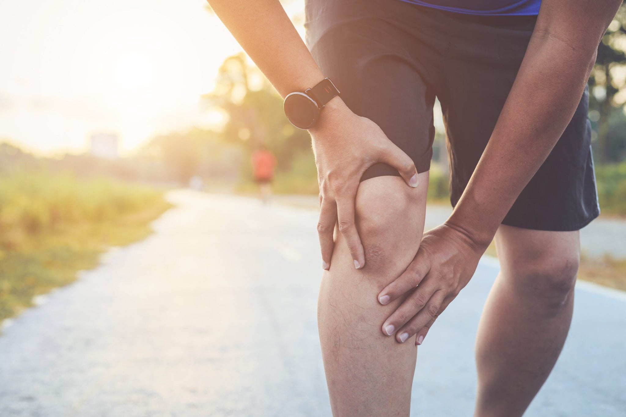 A person wearing a smartwatch is holding their knee in apparent discomfort while standing on an outdoor path.