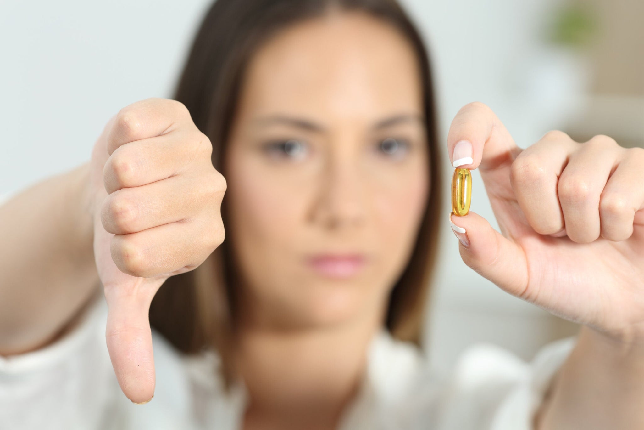 A woman giving a thumbs-down gesture while holding a yellow pill in her other hand. The background is blurred.