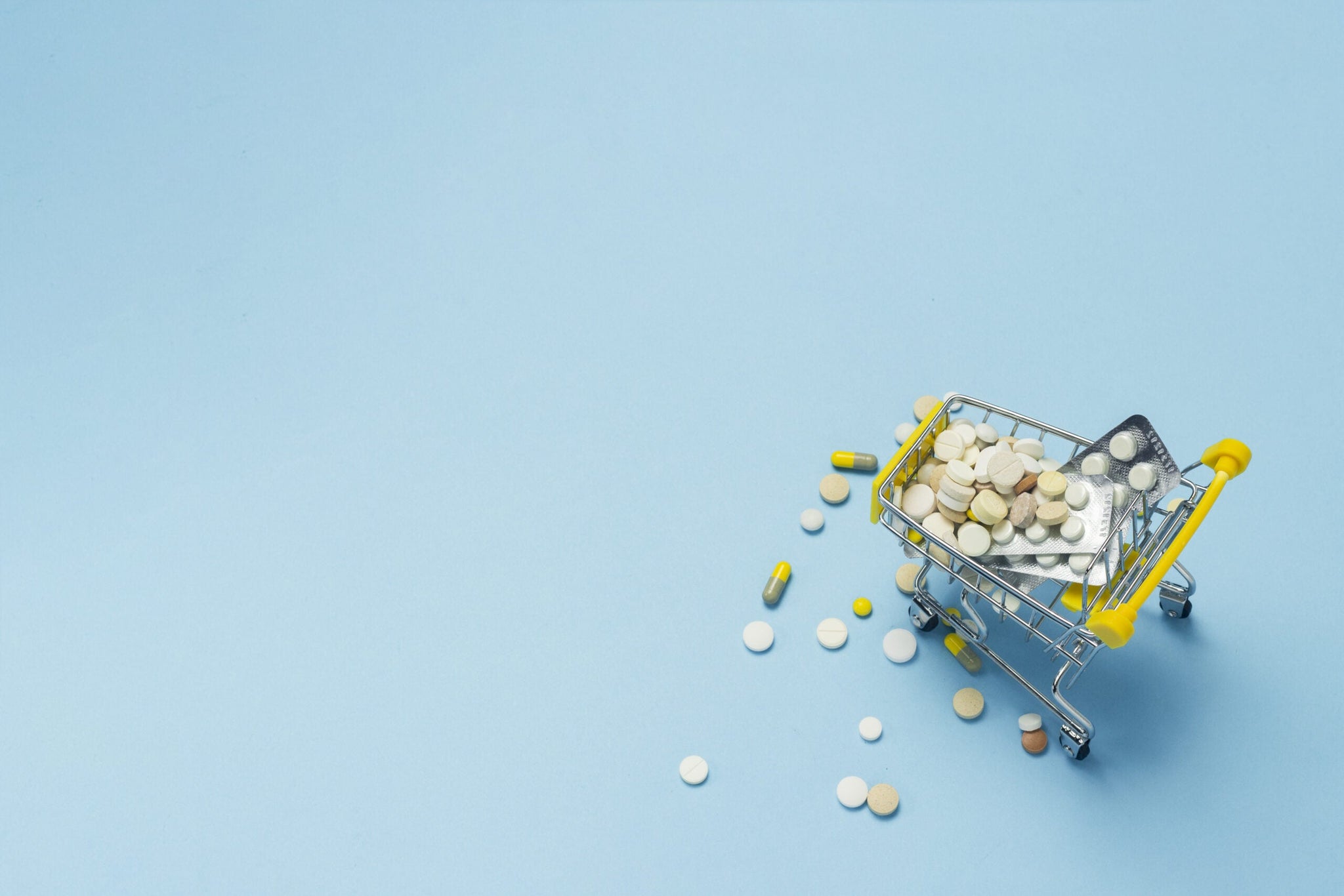 A small shopping cart filled with various pills and tablets, some of which are scattered around it against a light blue background.
