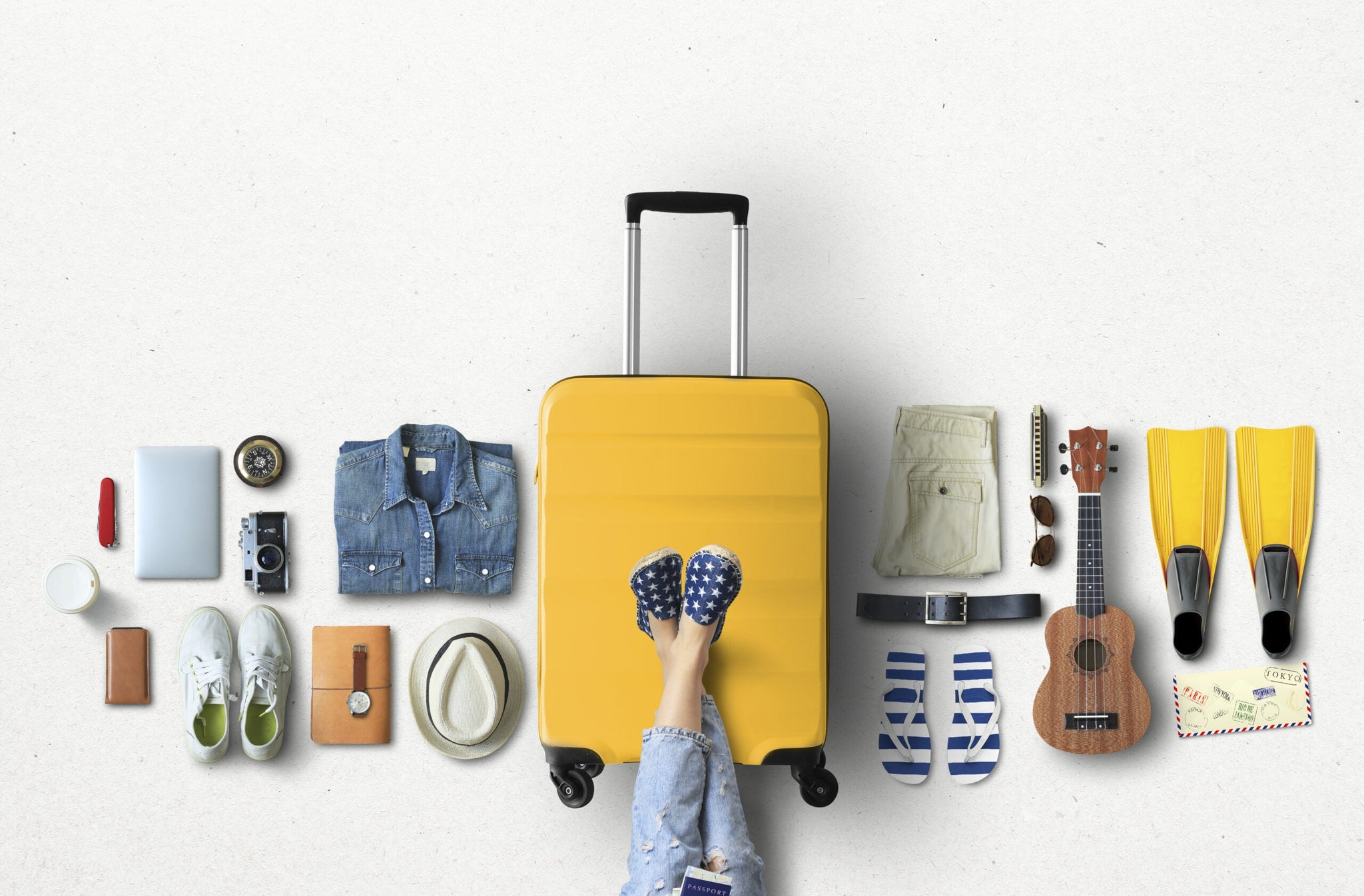 Overhead view of various travel items neatly arranged around a central yellow suitcase.