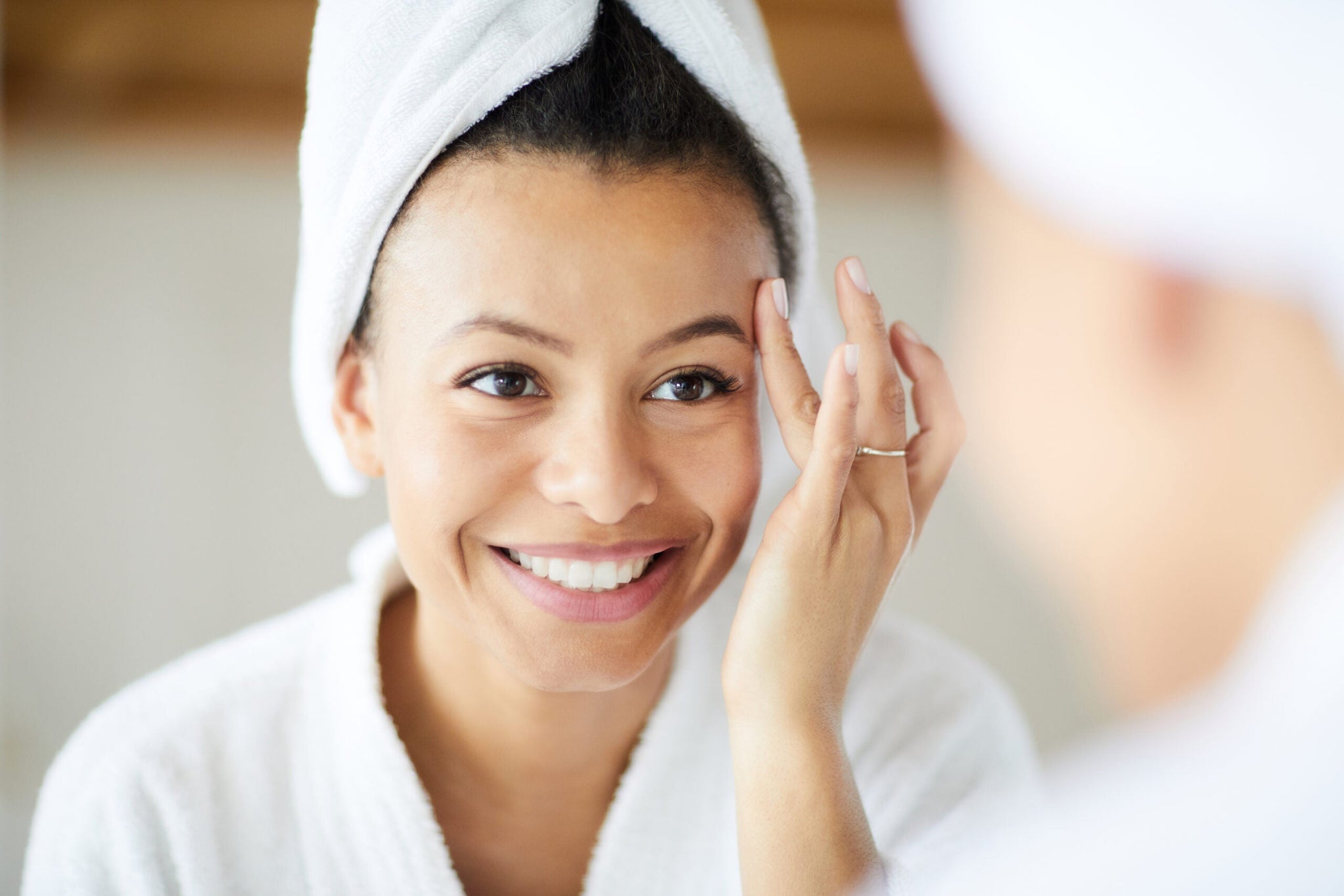 A smiling woman with a towel wrapped around her head stands in front of a mirror, touching her face gently.