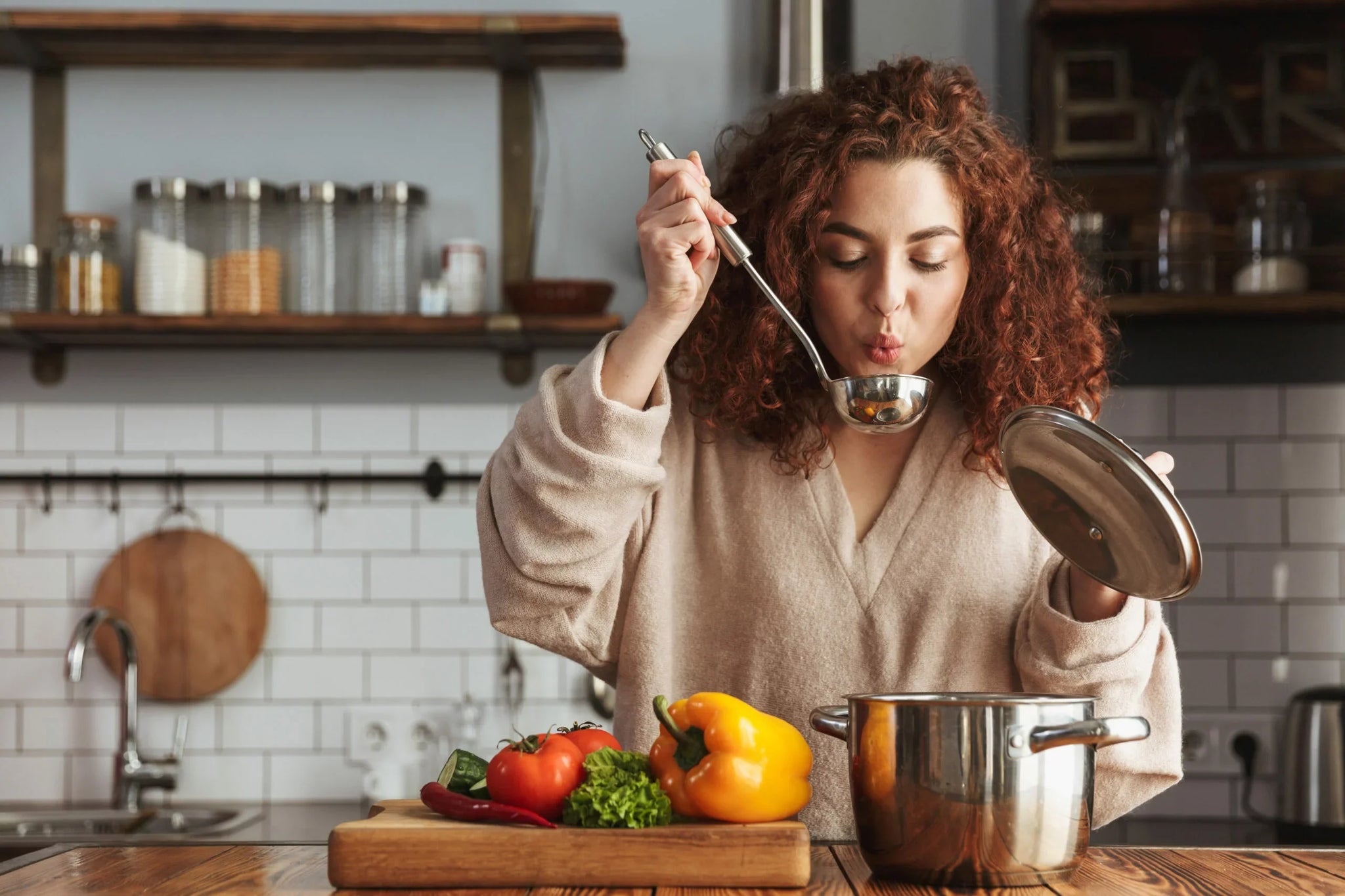 A woman with curly red hair wearing a beige sweater is blowing on a ladle filled with soup over a pot in a kitchen.