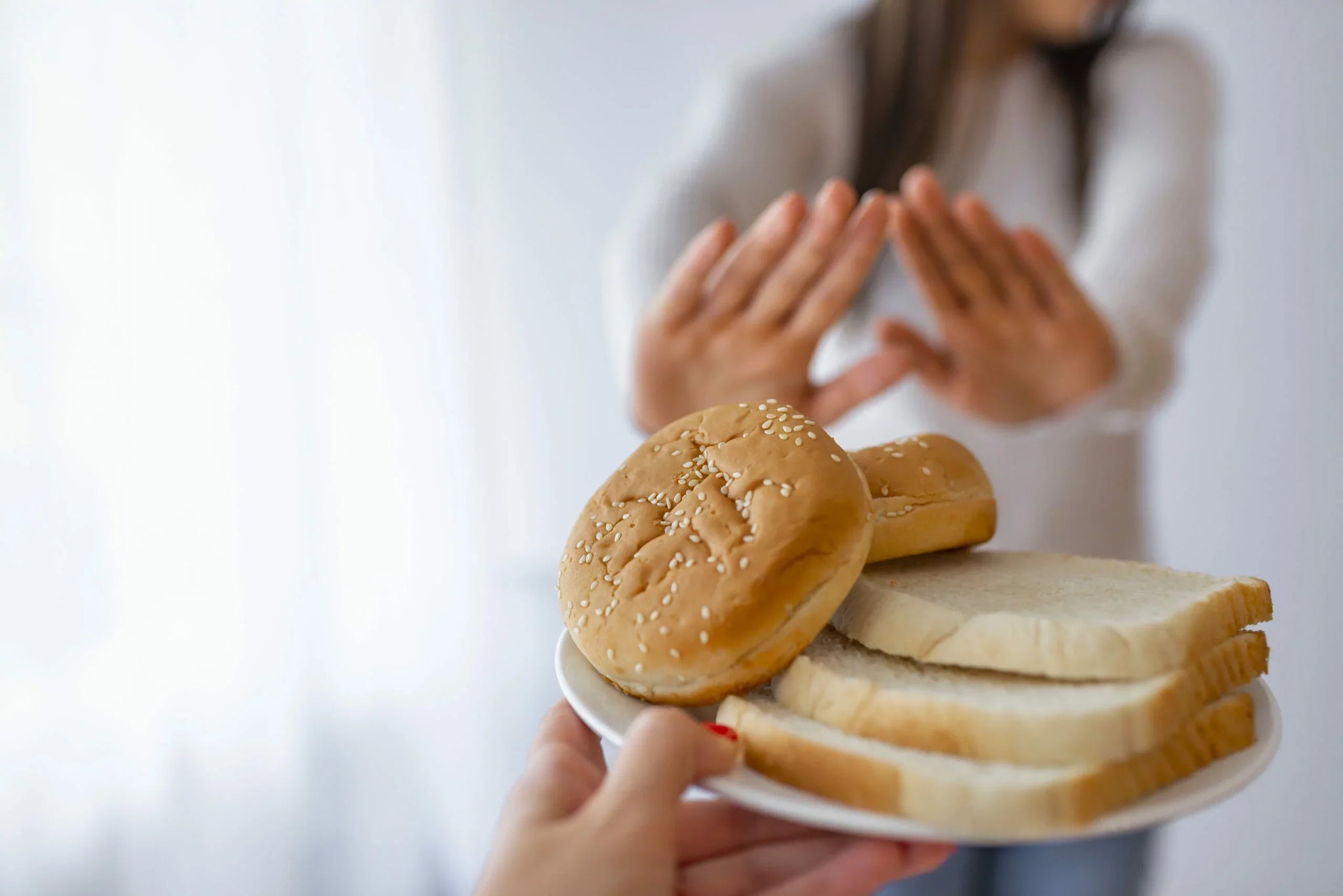 A person holds a plate with hamburger buns and slices of white bread in the foreground, offering it to a blurred figure in the background.