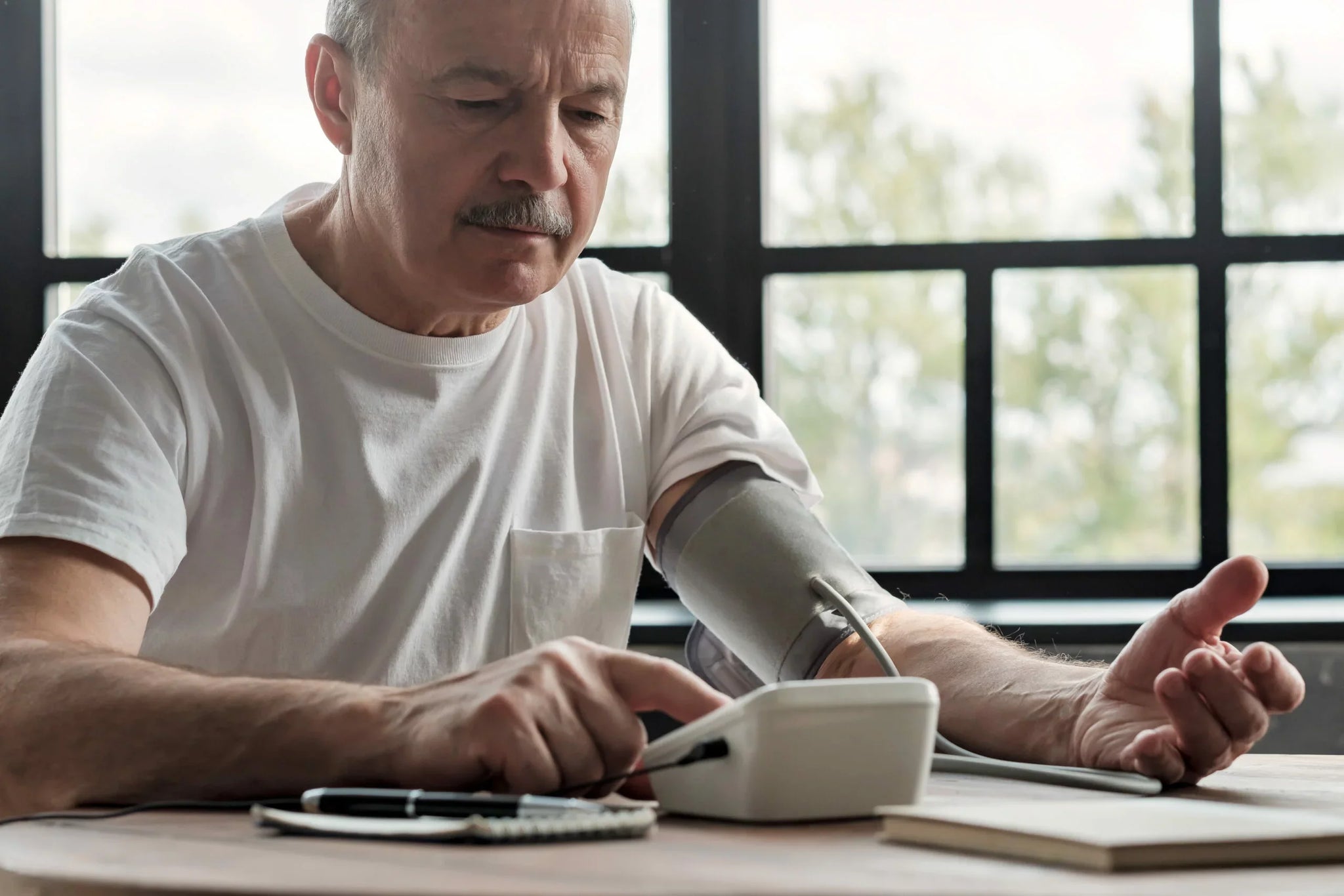 A man wearing a white t-shirt sits at a table by a large window, using a digital blood pressure monitor on his left arm.