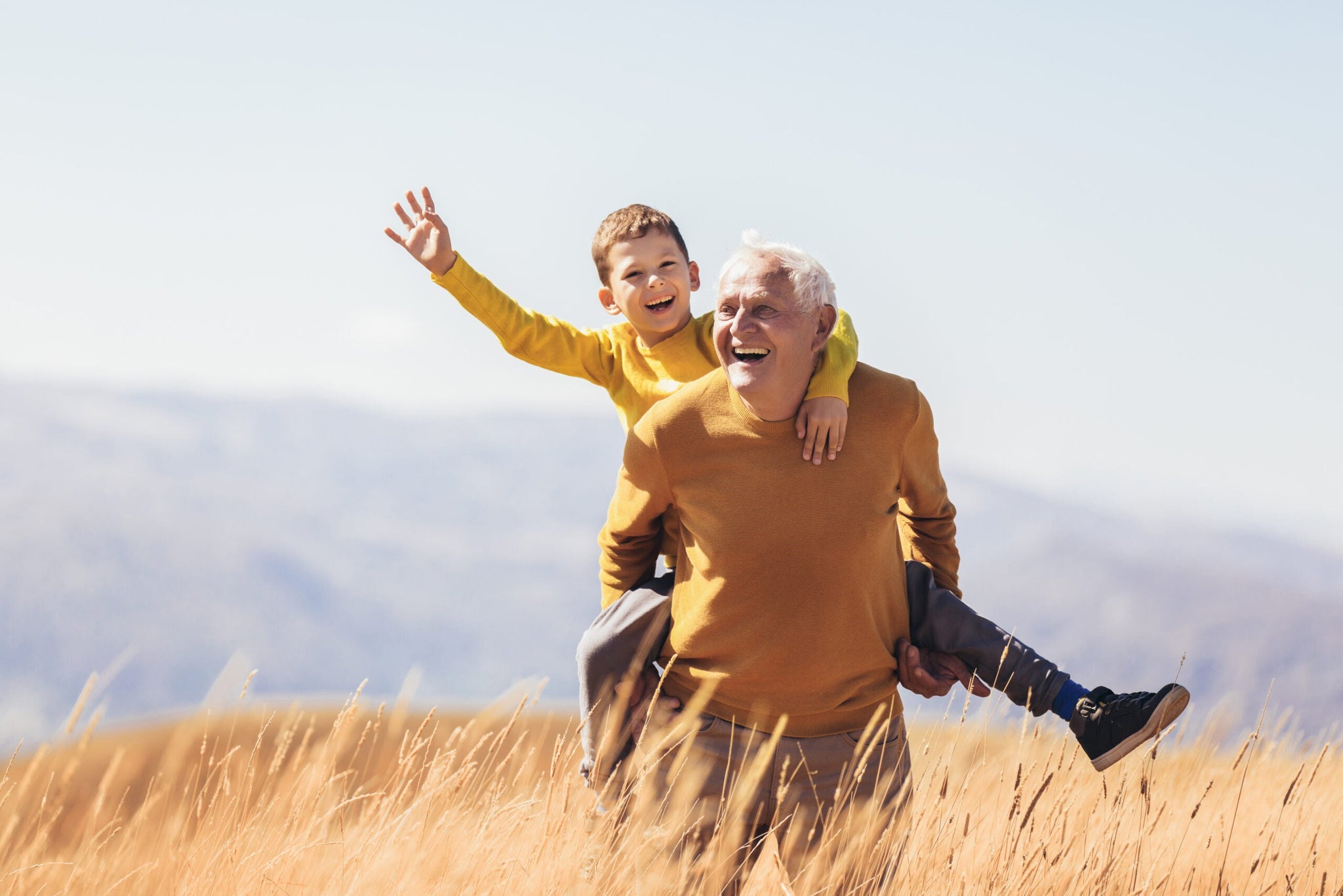 A joyful elderly man in an orange sweater gives a piggyback ride to a young boy in a matching orange sweater.
