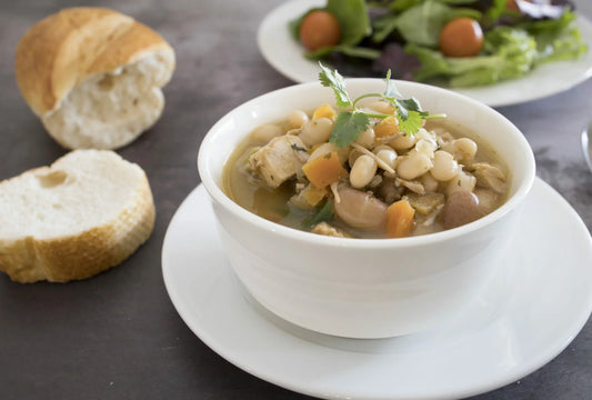 A bowl of white bean and vegetable stew garnished with cilantro, served on a white plate next to a piece of sliced bread.