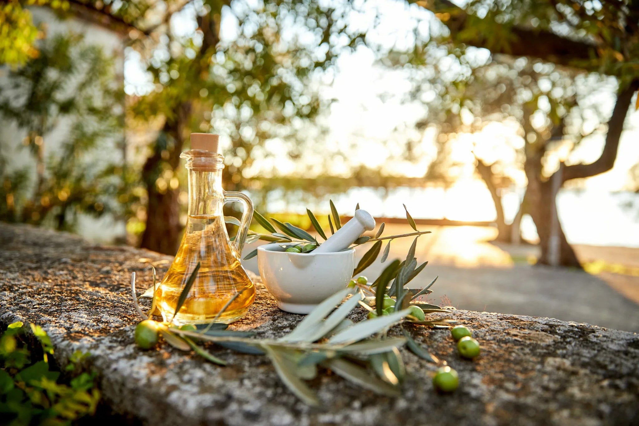 A glass bottle of olive oil and a white mortar and pestle filled with olive branches and leaves sit on a stone surface outside.