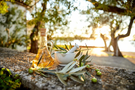 A glass bottle of olive oil and a white mortar and pestle filled with olive branches and leaves sit on a stone surface outside.