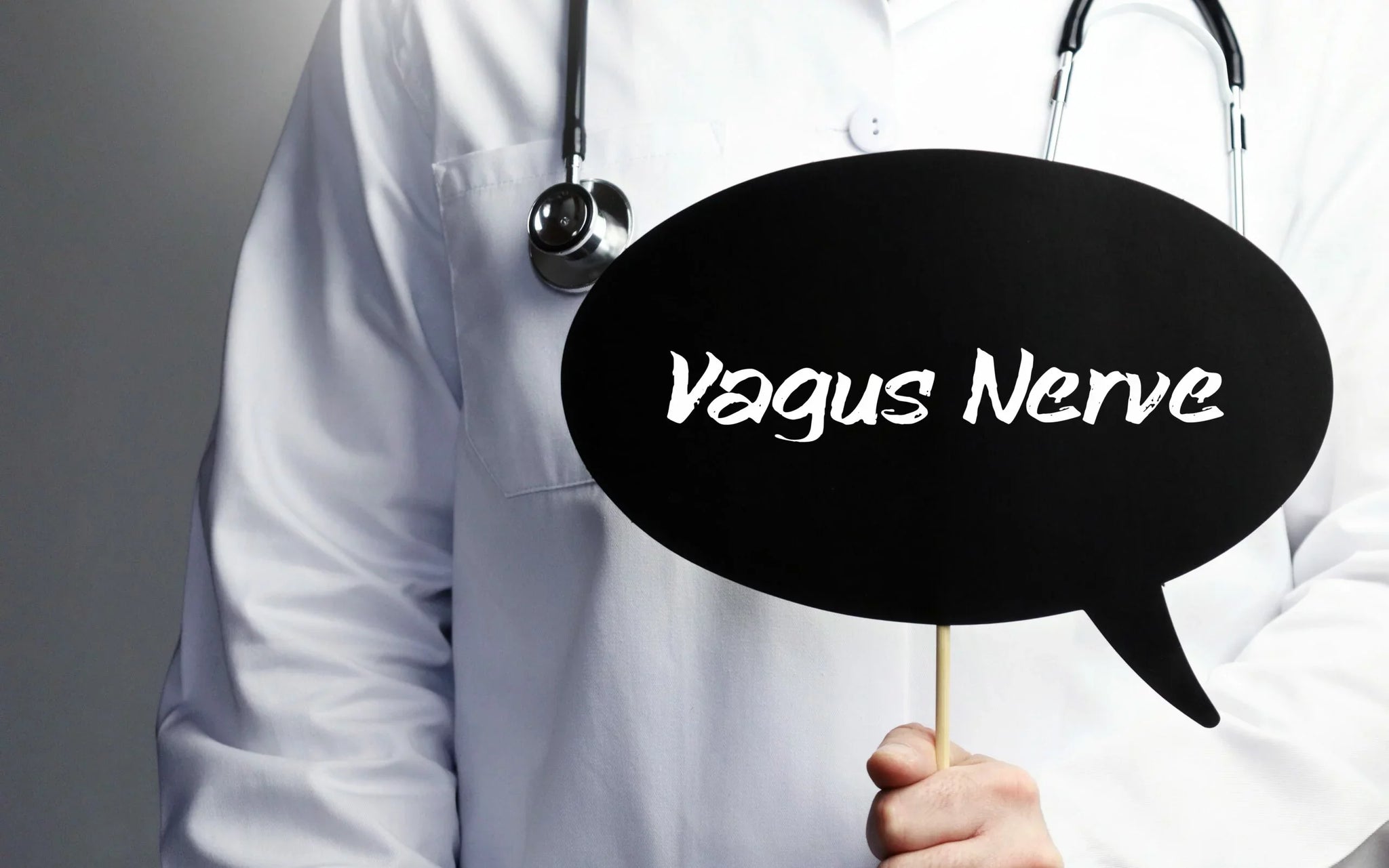 A person in a white lab coat, with a stethoscope draped around their neck, holds a black speech bubble sign with the words 