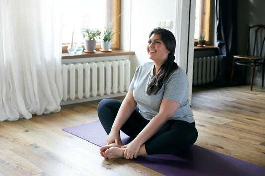 A woman sits on a purple yoga mat in a butterfly stretch position, smiling and looking relaxed.