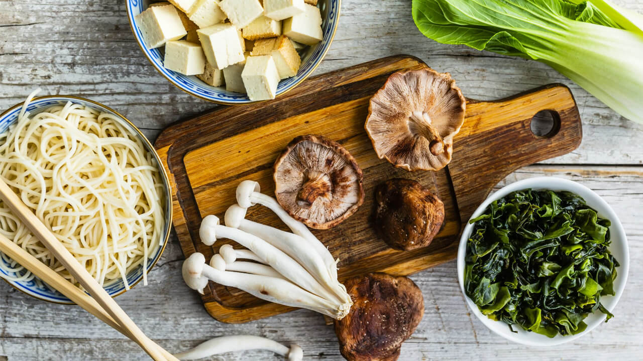 A wooden cutting board with assorted mushrooms, tofu cubes, and bok choy next to bowls of noodles, leafy greens