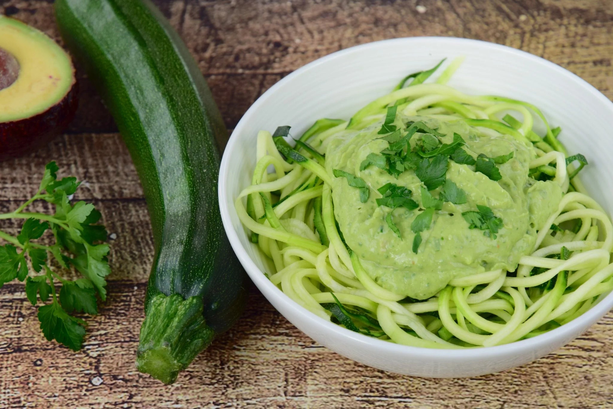 A white bowl filled with zucchini noodles topped with a creamy green sauce and garnished with fresh herbs.