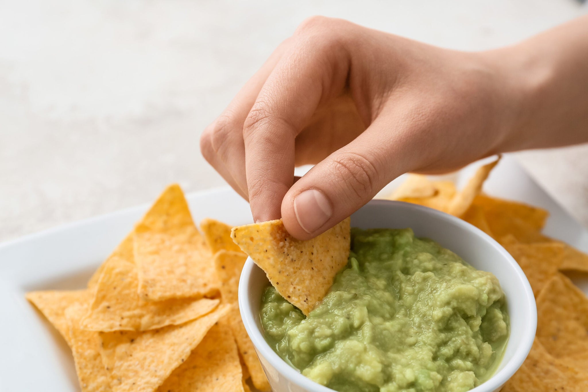 A hand is dipping a triangular tortilla chip into a bowl of creamy green guacamole.