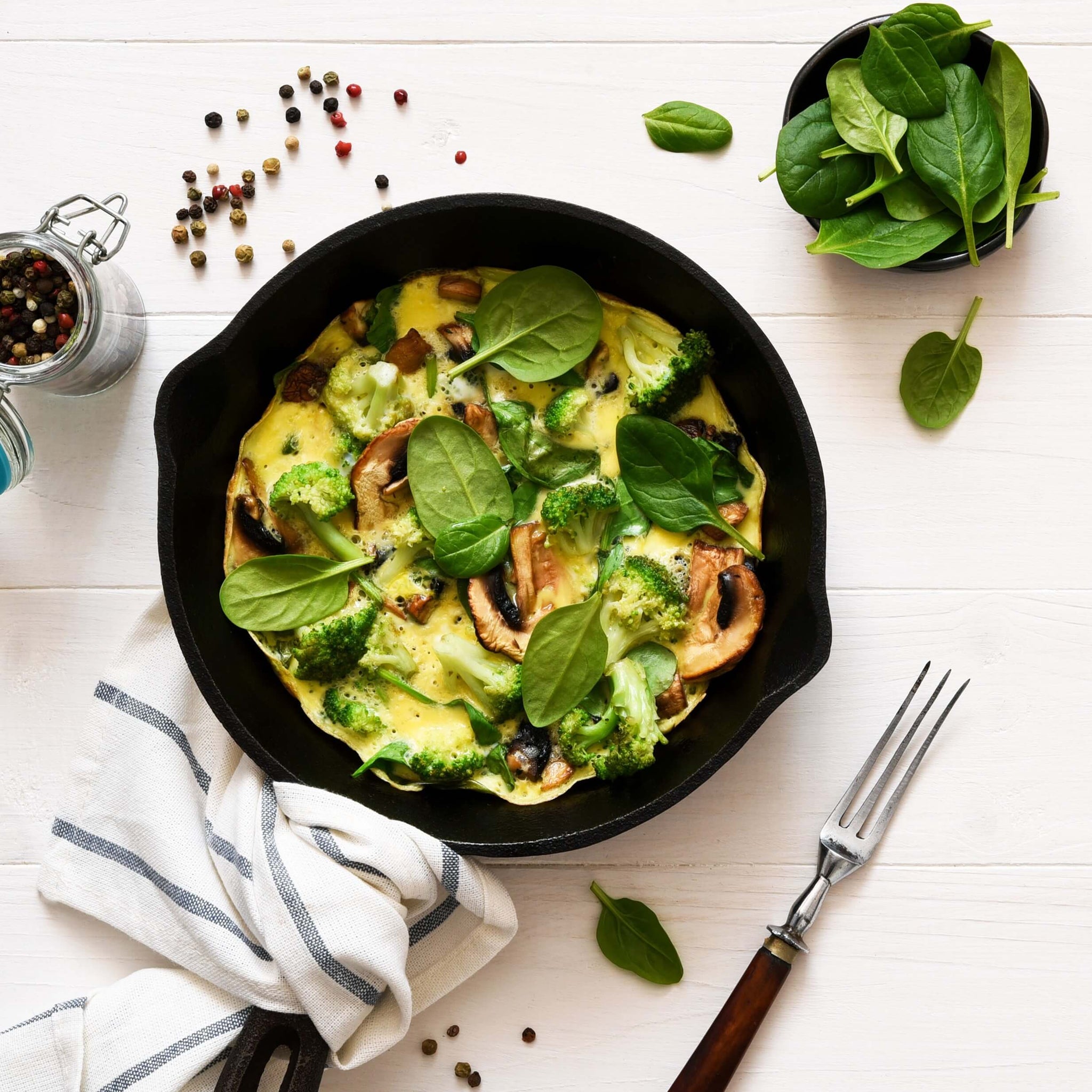 A cast-iron skillet filled with an omelette that includes mushrooms, broccoli, and fresh spinach leaves.