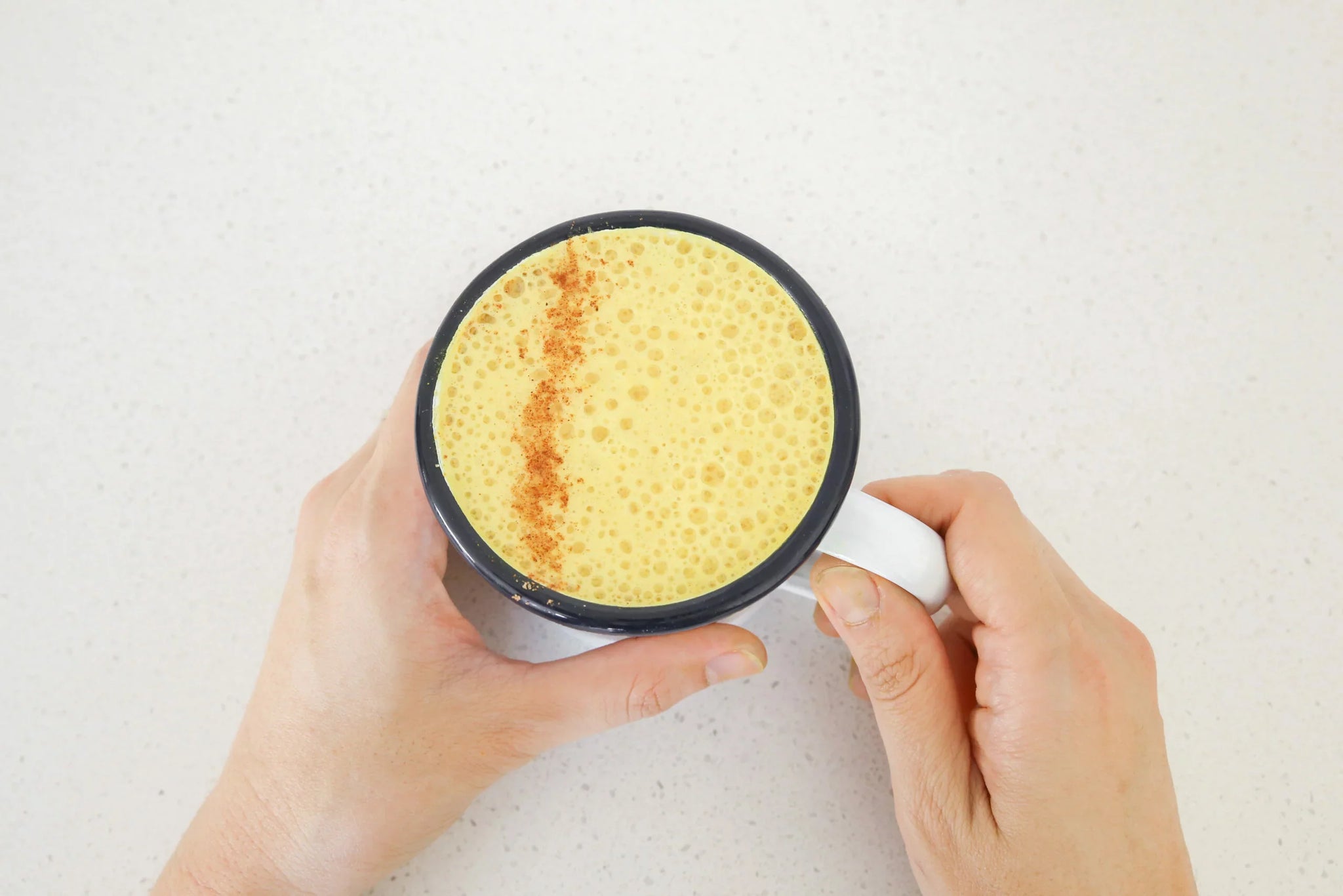 A person holding a white mug filled with a frothy yellow beverage, possibly turmeric latte, with a sprinkle of cinnamon on top.