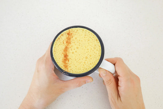 A person holding a white mug filled with a frothy yellow beverage, possibly turmeric latte, with a sprinkle of cinnamon on top.