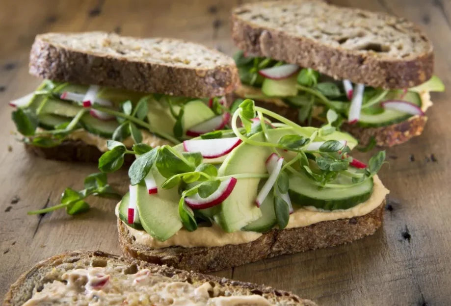 Three sandwiches made with whole grain bread, hummus, and fresh vegetables including cucumbers, radishes, and green sprouts
