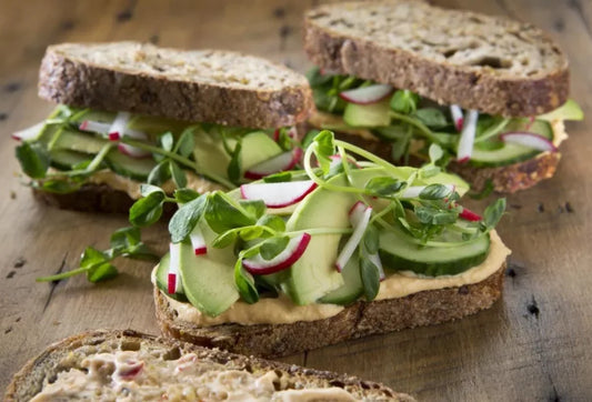 Three sandwiches made with whole grain bread, hummus, and fresh vegetables including cucumbers, radishes, and green sprouts