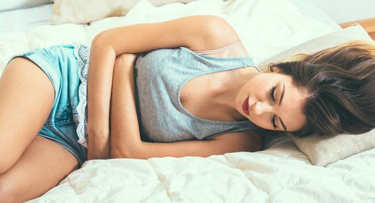 A woman lies on her side on a bed, clutching her stomach with both hands, clearly experiencing menstrual cramps.