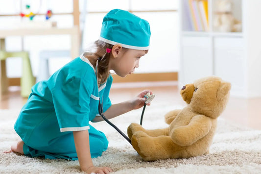 A young child dressed in teal medical scrubs and a matching cap kneels on a carpeted floor using a stethoscope to check a teddy bear.