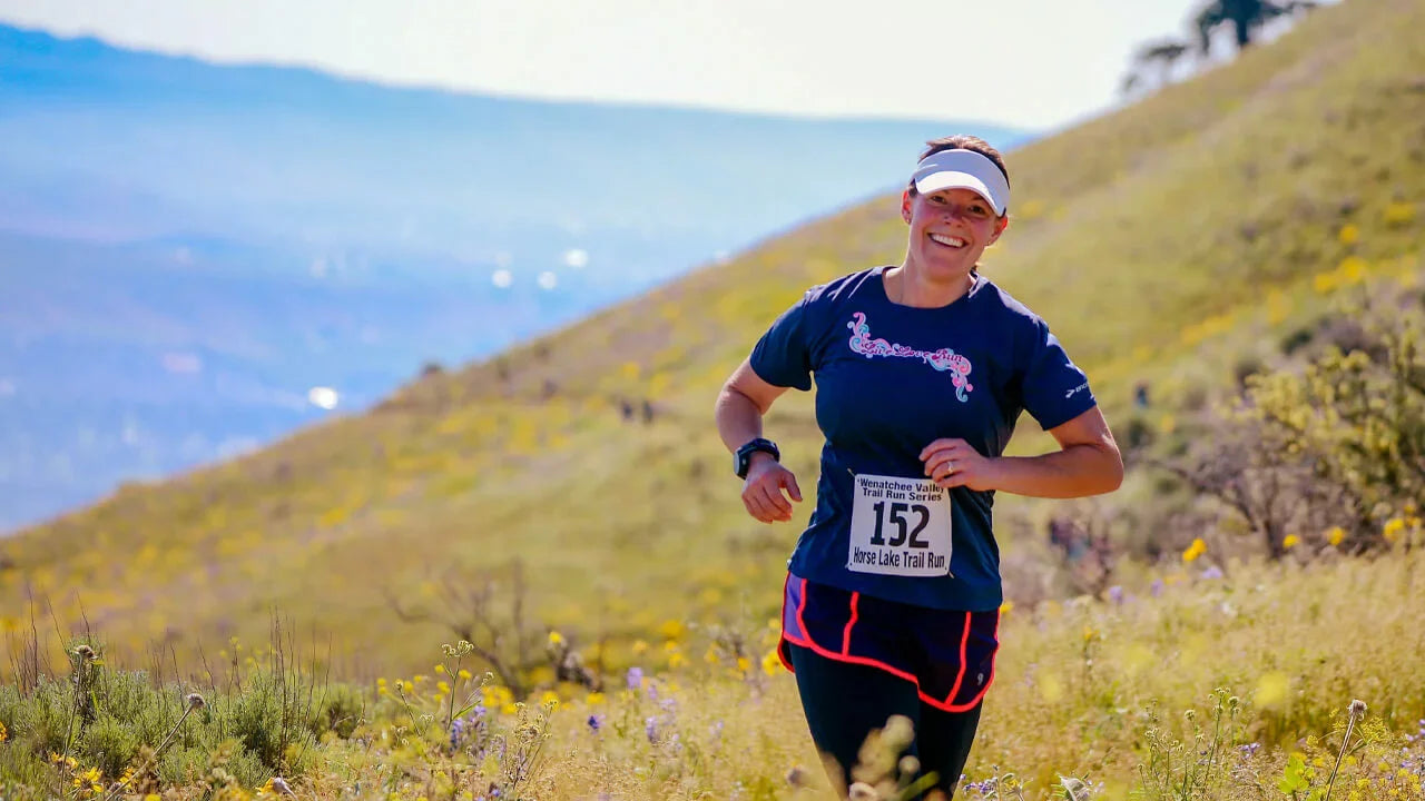 A person wearing a blue shirt, black shorts, and a white visor runs on a grassy hill with race number 152 on their chest,