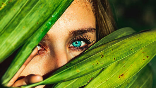 A close-up of a woman's face partially obscured by large green leaves. One blue eye, benefiting from omega-3, is visible, looking directly at the camera and framed by dark eyelashes.
