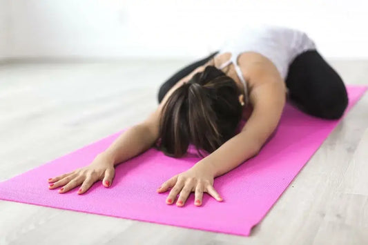 A woman is performing a yoga child's pose on a bright pink yoga mat in a spacious room.