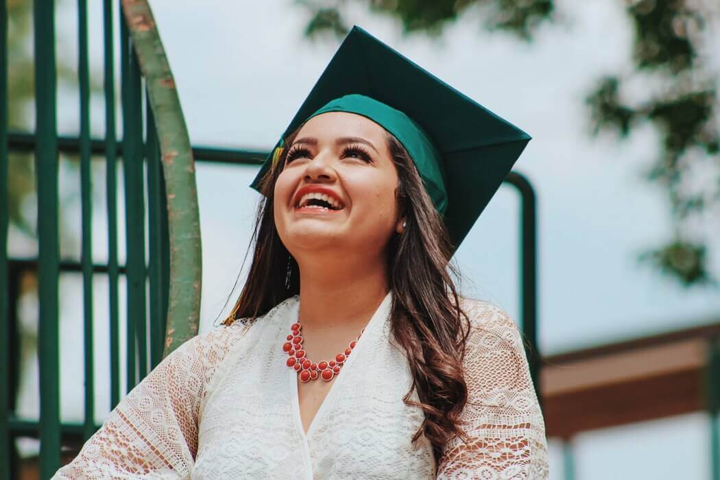 A 3 woman in a green graduation cap and white lace dress smiles brightly while looking up, celebrating the added benefits series of her achievements.