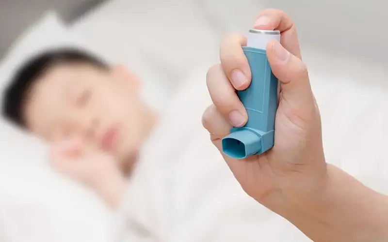 A hand holding a blue inhaler in the foreground, with a blurred background showing a person lying down, possibly sleeping, on a bed.