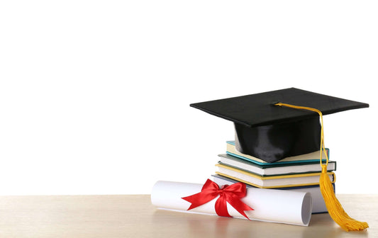 A graduation cap with a yellow tassel sits on top of a stack of books next to a rolled-up diploma tied with a red ribbon