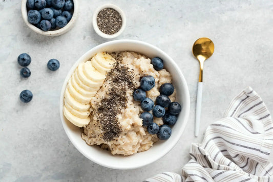 A bowl of oatmeal topped with sliced banana, blueberries, and chia seeds is on a light gray surface.