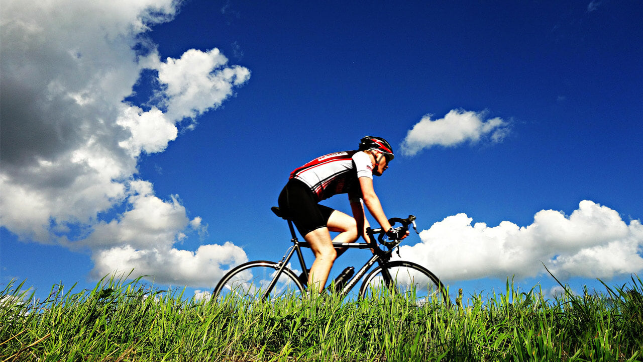 A cyclist in a black and white outfit and helmet rides a road bike on a path lined with green grass. The sky above is bright blue with scattered white clouds, making it a perfect day for pondering, 