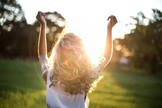 A person with long, wavy hair stands outside with their arms raised towards the sky, facing the sun.