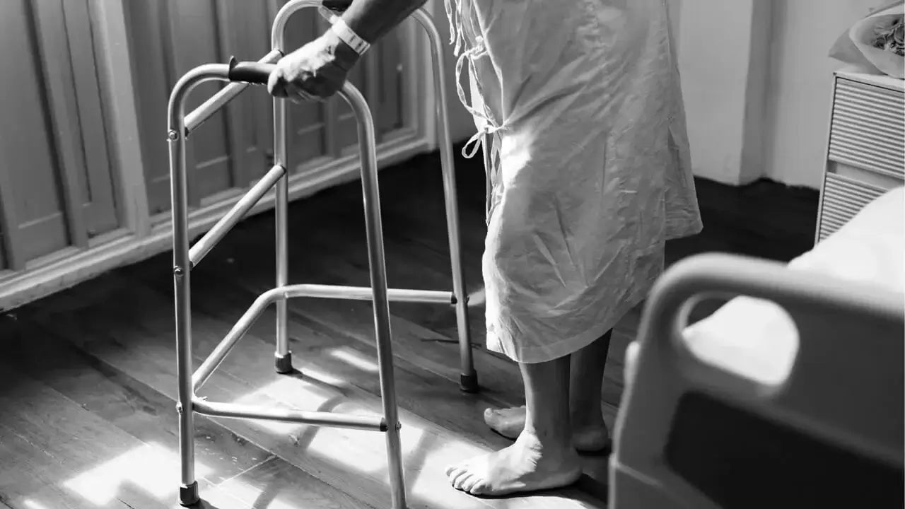 A person in a hospital gown, possibly experiencing early lupus symptoms, uses a walker for support while standing on a wooden floor.