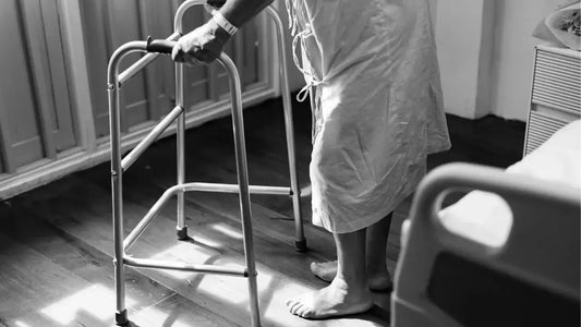 A person in a hospital gown, possibly experiencing early lupus symptoms, uses a walker for support while standing on a wooden floor.