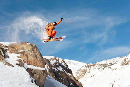 A skier in an orange outfit performs a jump off a rocky ledge on a snowy mountain, with snow spraying around them. Action sports