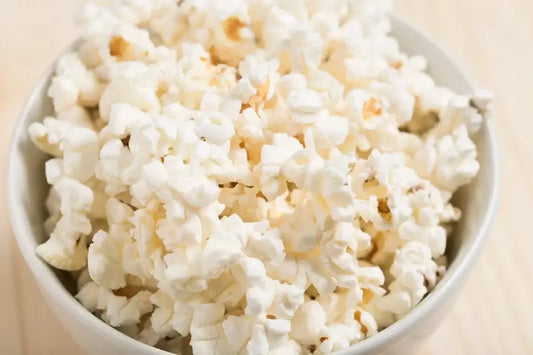 A white bowl filled to the brim with freshly popped popcorn sits on a light wood surface.